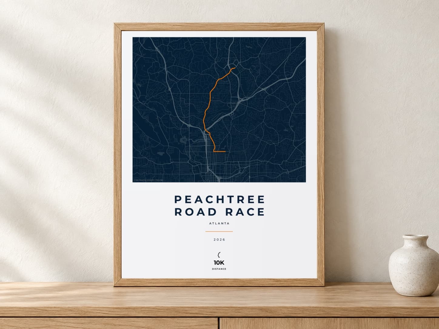 Peachtree Road Race 2026 Race Blueprint route print in a light oak frame on a console