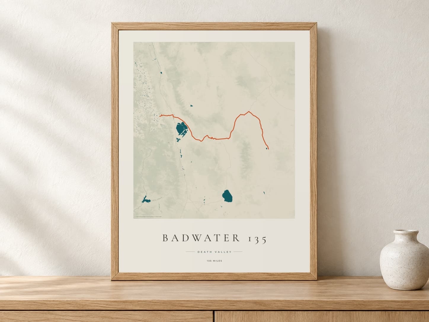 Badwater 135 2026 Coastal Atlas route print in a light oak frame on a console